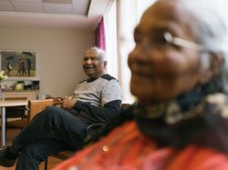 Een oudere man zit in een stoel met zijn handen in elkaar gevouwen en lacht breed.