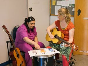 Persoon geeft gitaarles aan persoon met donker haar in twee staartjes. De persoon wijst naar een afbeelding van iemand die blij is