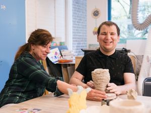 Een mijnheer met zwarte polo maakt een pot met stukken klei. Hij kijk blij naar de camera. Een begeleider met lang roodbruin haar kijkt naar zijn werk