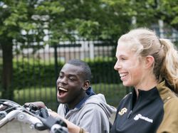 Jonge man met kort zwart haat zit met een grote lach naast een begeleider met blonde paardenstaart p een duo-fiets.