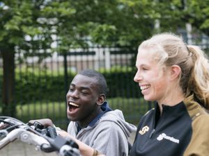 Jonge man met kort zwart haat zit met een grote lach naast een begeleider met blonde paardenstaart p een duo-fiets.