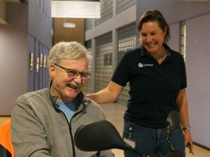 Medewerker met blauwe Cordaan-polo staat naast oudere meneer in scootmobiel met hand op zijn schouder en ze lachen
