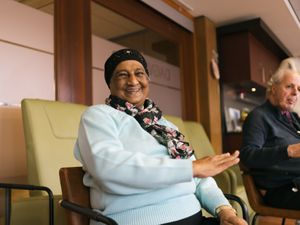 Oudere mevrouw met lichtblauwe trui, bloemensjaal en zwarte haarband zit in een stoel en zwaait met haar arm.