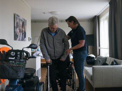 Medewerker met blauwe Cordaan-polo helpt oudere meneer van een rolstoel in een scootmobiel