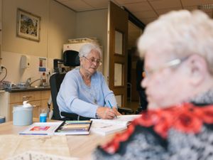 Dame met kort grijs haar in rolstoel zit met andere dame met grijs haar en bril aan tafel. Zij kleurt in een kleurboek, op tafel een doos met tientallen potloden.