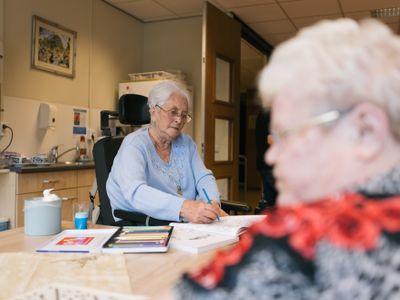 Dame met kort grijs haar in rolstoel zit met andere dame met grijs haar en bril aan tafel. Zij kleurt in een kleurboek, op tafel een doos met tientallen potloden.