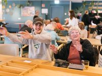 Een jongeman met een pet en een oudere damen zitten aan tafel voor een sjoelbak en zwaaien mee met muziek