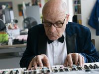 Oudere heer met bril en donkerblauw colbert, wit overhemd en strik kijkt geconcentreerd naar de keyboard waar zijn handen op rusten.