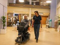 Medewerker met blauwe Cordaan-polo loopt naast oudere meneer in scootmobiel door de hal