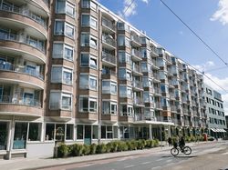 Gebouw van 6 verdiepingen, met in het midden de ingang. Op de voorgrond een lage, groene heg en een fietser die de weg oversteekt.