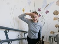 Meisje met grijs shirt en rode nagellak loopt een trap op en kijkt met haar hand tegen haar voorhoofd naar boven. De muur is behangen met grote bloemen en insecten.