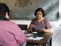 Twee oudere dames zitten aan tafel en spelen een kaartspel. Aan de wand hangt een groot kleed.