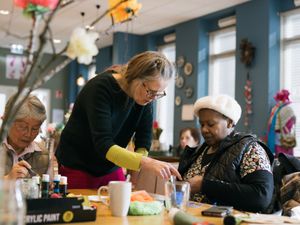 Vrouwelijke begeleider kijkt aandachtig naar het handwerk van een dame die aan tafel werkt