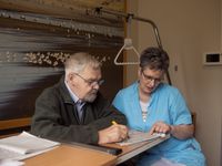 Mijnheer met baard en bril zit aan een tafel en vult een lijst in samen met een verzorgende met bril in blauw uniform