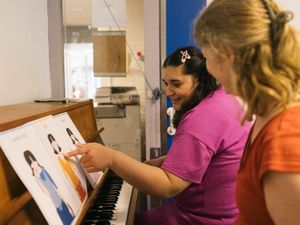 Persoon geeft pianoles aan persoon met donker haar. De persoon wijst in een boekje het plaatje dat voor blij staat aan
