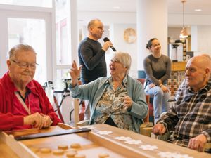 Drie vrolijke oudere mensen zitten aan tafel bij een sjoelbak en achter hen zingt een man met microfoon