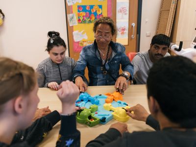 Een begeleider speelt een spelletje aan tafel met cliënten aan tafel. Op de achtergrond hangen vrolijke tekeningen aan de deur.
