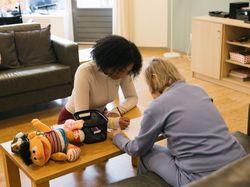 1 persoon helpt een ander persoon om een tekening te maken aan de salontafel in de woonkamer. Knuffel Ernie ligt er bij