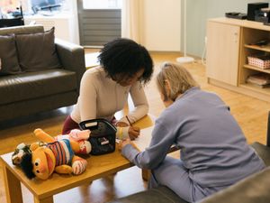1 persoon helpt een ander persoon om een tekening te maken aan de salontafel in de woonkamer. Knuffel Ernie ligt er bij