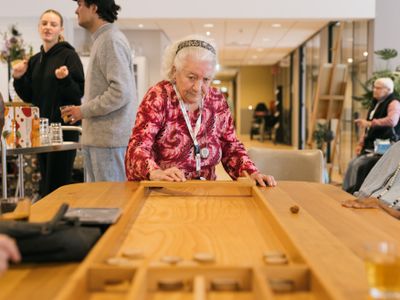 Een oudere dame met een truitje met drukke roze print en een panterprint haarband is aan het sjoelen in een activiteitenruimte
