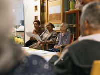 Drie vrouwen zitten naast elkaar te zingen op een stoel. Ze hebben blaadjes vast met daarop de songtekst.