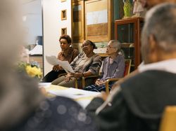 Drie vrouwen zitten naast elkaar te zingen op een stoel. Ze hebben blaadjes vast met daarop de songtekst.