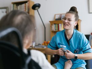 Een lachende medewerker helpt iemand die in een rolstoel zit, met sokken aantrekken. Op de achtergrond staan boekenkasten en een eettafel.