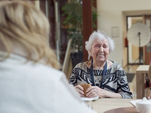 Dame met grijs haar zit aan tafel met een tafelgenoot en lijkt lachend in de camera met een bruin broodje in haar hand