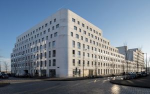Een groot, wit, modern gebouw met in de lengte verschillende aantallen verdiepingen en kleine ramen, aan de hoek van een straat.