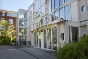 Moderne gebouwgevel met een glazen entree en een rond "Vivaldi"-logo, omgeven door groen en aangrenzende gebouwen.