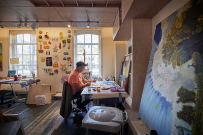 Een man met oranje shirt en pet zit in een ruimte vol schilderijen en maakt zelf ook een kleurrijk schilderij.