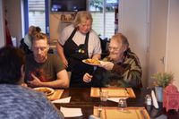 Drie mensen zitten aan een gedekte tafel. Een vrouw met schort met kleurrijk SamSam logo geeft aan één van hen een bord met eten.