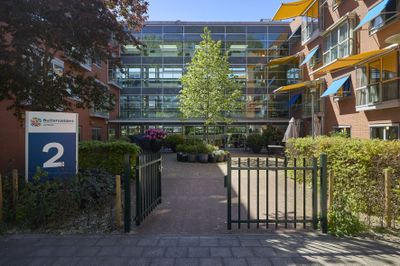 Ingang van een modern gebouw met glazen gevel, groen, zitgelegenheid buiten en kleurrijke driehoekige zonneschermen onder een helderblauwe hemel.