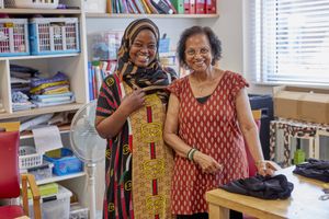 Twee donkere vrouwen lachen vriendelijk naar de camera terwijl ze creatief bezig zijn in de textielwerkplaats van Osdorperhof.
