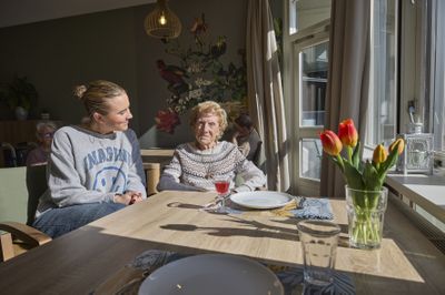 Een oude vrouw en een jonge vrouw zitten lachend naast elkaar aan een houten tafel in een zonnige ruimte. De tafel is gedekt.