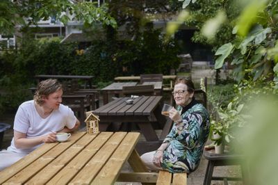 Een vrolijk gesprek tussen een mannelijke begeleider en een oudere deelneemster aan de dagbesteding. De begeleider drinkt een soepje en de vrouw een koffie. Ze zitten gezellig samen buiten aan een houten picknicktafel.