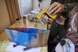 Een vrouw spuit met hulp van iemand blauwe verf op een kartonnen doos.