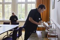 Een man en een vrouw met zwarte kleding zetten koffiekopjes neer in een zaal met een grote tafel en stoelen.