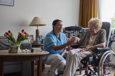 Een bewoonster in een rolstoel krijgt haar medicatie aangereikt van een medewerkster in een blauw uniform.
