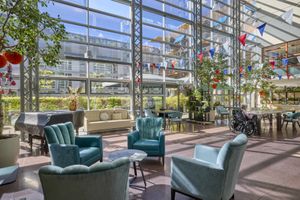 Een zonovergoten atrium met turquoise fauteuils, feestelijke slingers, planten en grote ramen die uitzicht bieden op een modern stadslandschap.