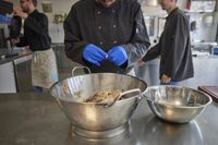 In een industriële keuken werkt iemand met een zwarte koksbuis en blauwe handschoenen aan. Hij bereidt kip in een grote vergiet. Op de achtergrond zie je 2 personen in dezelfde werkkleding.