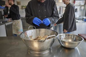 In een industriële keuken werkt iemand met een zwarte koksbuis en blauwe handschoenen aan. Hij bereidt kip in een grote vergiet. Op de achtergrond zie je 2 personen in dezelfde werkkleding.