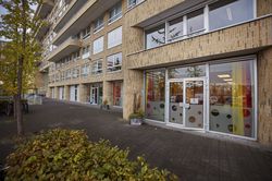 Gebouw met een bakstenen gevel met balkons en veel ramen. Op de begane grond staat een deur open. Er hangen gordijnen in rood, geel en oranje. Voor de deur is een brede stoep met bomen en een fietsrek.