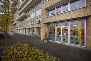 Gebouw met een bakstenen gevel met balkons en veel ramen. Op de begane grond staat een deur open. Er hangen gordijnen in rood, geel en oranje. Voor de deur is een brede stoep met bomen en een fietsrek.