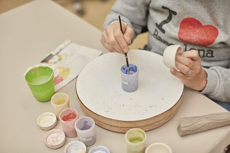 Iemand in een grijze trui decoreert een schaaltje van keramiek met blauwe glazuur. Op de voorgrond staan potjes met verschillende, vrolijke kleuren glazuur.