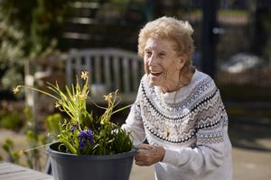 Een oudere dame lacht breed terwijl ze een bak met bloemen optilt.