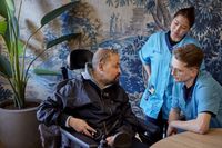 Naast een man in een rolstoel staan twee jonge zorgmedewerkers in blauwe uniformen. Ze zijn met elkaar in gesprek.