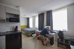 Een man zit achter zijn bureau met computer en muziekinstallatie in een woning met geel bed en grijze gordijnen.