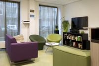 Een kamer met groene banken en stoelen, een tv aan de muur en daaronder een zwarte kast met vakken met boeken.