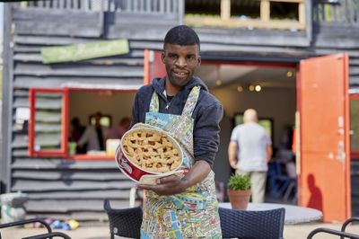 Een man met een schort bedrukt met plattegrond van Amsterdam staat voor een buitencafé en houdt trots een verse appeltaart in bakvorm vast.